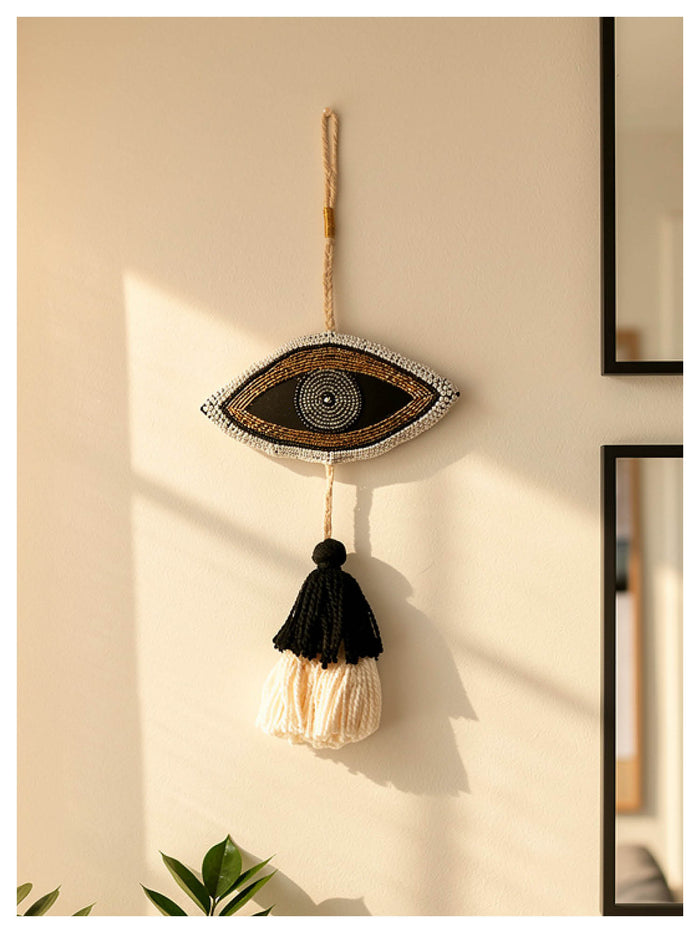 Decorative wall hanging with eye design and tassels on a beige wall.