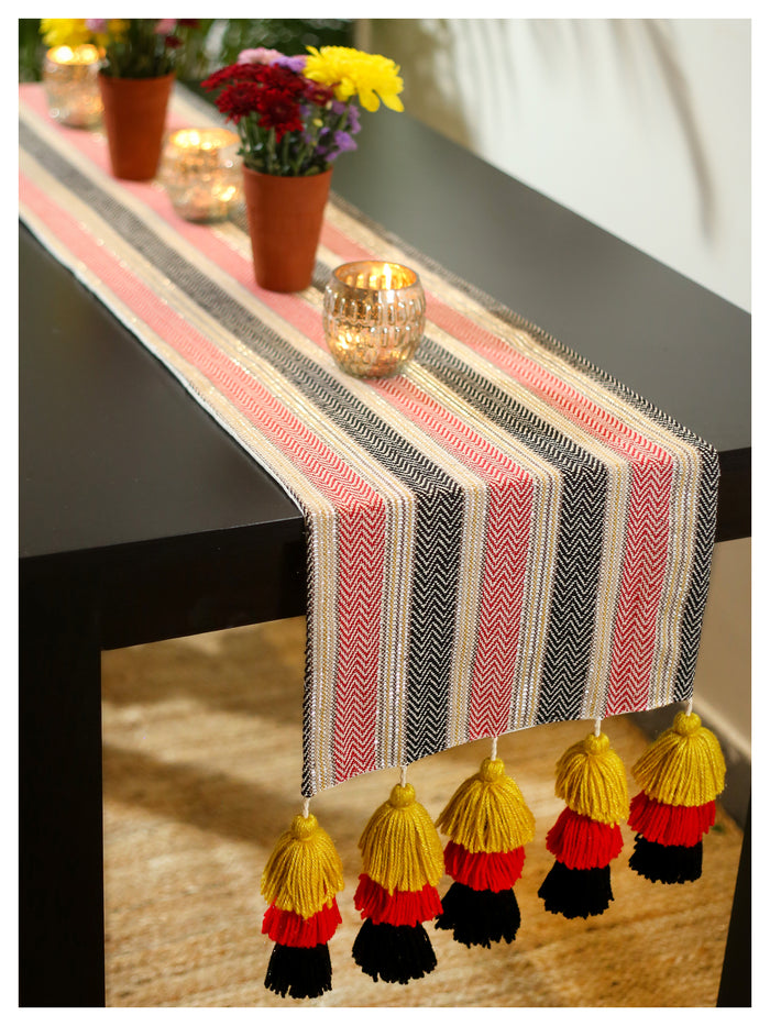 A table runner with a red mustard and black stripe pattern, featuring tassel detail, displayed on a table with potted plants and candles.